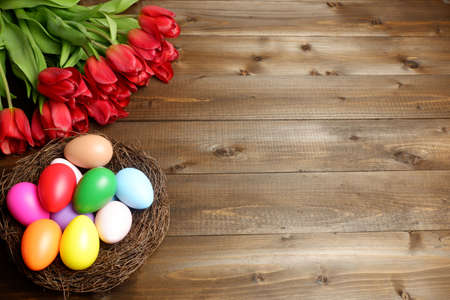 Birds nest with multicolored easter eggs and bouquet of red tulip flowers on dark wooden planks desk. Easter holiday banner, card, poster, header, voucher, invitation templateの写真素材