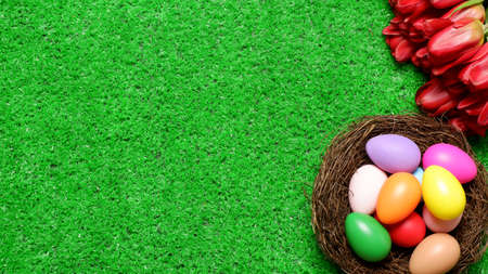 Multi colored easter eggs in birds nest and bouquet of red tulip flowers on green grass lawn background top view with copy space. Easter holiday banner, card, poster, voucher, header templateの写真素材