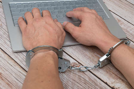 A man's hands in handcuffs and laptop on wooden table. Piracy, internet hacking and punishment for cybercrime concept.の写真素材