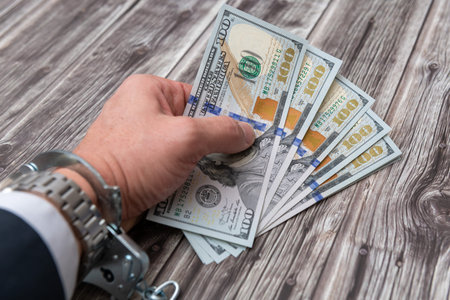 A man's hand in handcuffs and US dollars banknotes on wooden table. Corruption or economic crime...