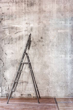 Home interior room repair concept - old gray concrete wall and dirty brown floor in repairing room with old dirty black stepladder near the wall closeup viewの写真素材