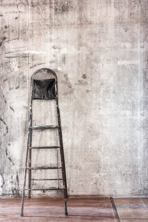 Home interior room repair concept - old gray concrete wall and dirty floor in repairing room with old dirty black stepladder near the wall closeup viewの写真素材
