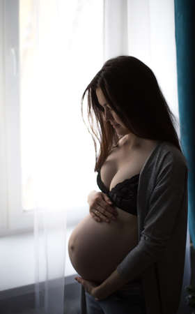 young pregnant girl keeps her belly and standing by the window. Waiting babyの写真素材