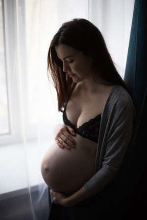 young pregnant girl keeps her belly and standing by the window. Waiting babyの写真素材