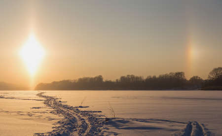A path in the snow retreating into the distance against the background of the winter landscape and the natural phenomenon of the parhelion or halo.の写真素材