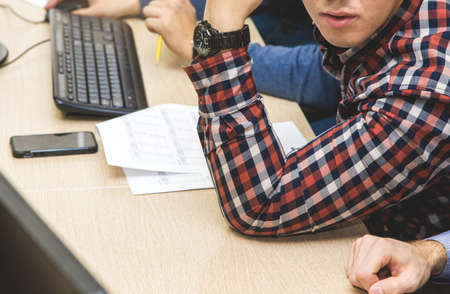 young man student in a checkered shirt at a table among papers and other office supplies studying or working in a computer room.の写真素材