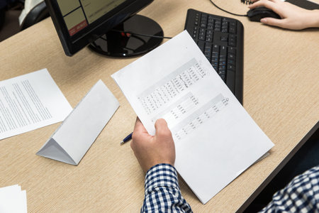 young man student in a checkered shirt at a table among papers and other office supplies studying or...