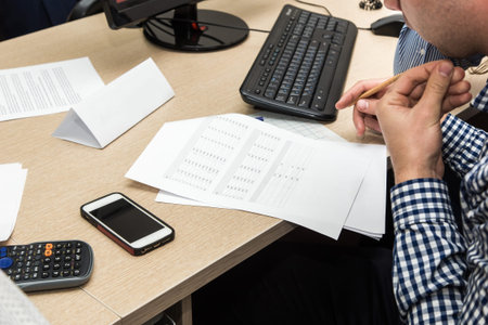 young man student in a checkered shirt at a table among papers and other office supplies studying or...