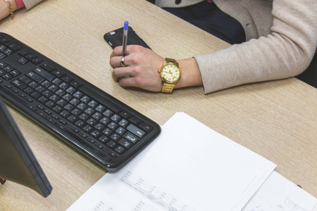Man is sitting at a desk by the computer among office supplies and is holding a pen in his hands with golden clock. Work in the office or studyの写真素材