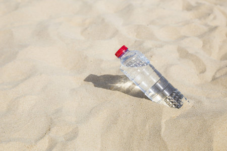 Bottle with crystal clear drinking water with a red lid on the sand on a hot, sunny day closeupの写真素材