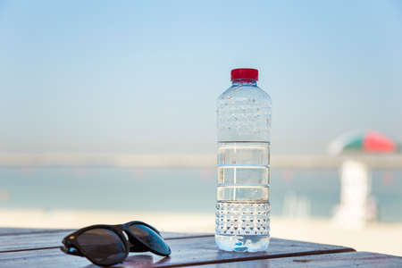 a plastic bottle with clean drinking water with a red lid on a wooden table and with sunglasses on the background of a palm and sea beach on a sunny summer dayの写真素材