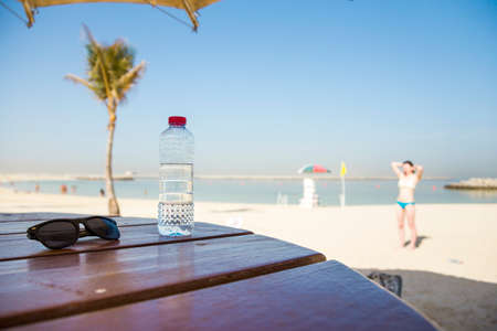 a plastic bottle with clean drinking water with a red lid on a wooden table and with sunglasses on the background of a palm and sea beach on a sunny summer dayの写真素材