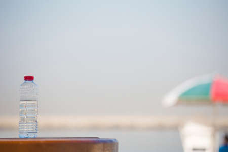 a plastic bottle with clean drinking water with a red lid on a wooden table against the background of the sea and beach umbrella on a sunny summer dayの写真素材