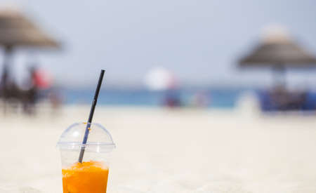 bright orange cold refreshing smoothie drink in a plastic cup with a black straw in the sand on a beach background on a hot sunny day. Summer conceptの写真素材