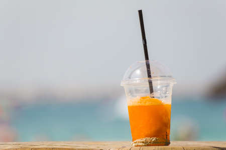 bright orange cold refreshing smoothie drink in a plastic cup with a black straw in the sand on a beach background on a hot sunny day. Travel conceptの写真素材