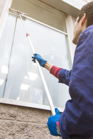 A young male cleaner in blue overalls washes windows and store front. Cleaning Service