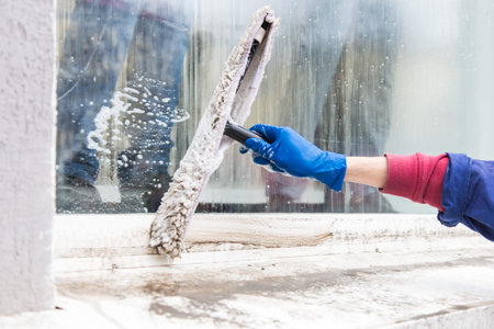 A young male cleaner in blue overalls washes building's facade, windows and store front. Cleaning...