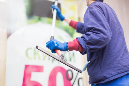 A young male cleaner in blue overalls washes windows and store front. Cleaning Service. inscription:...