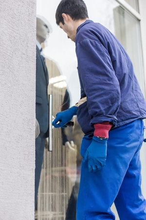 A young male cleaner in blue overalls washes windows and store front. Cleaning Service