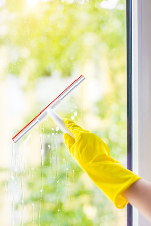 hand cleaning glass window pane with detergent and rubber aluminum wiper. A female hands in bright...