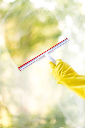 hand cleaning glass window pane with detergent and rubber aluminum wiper. A female hands in bright yellow gloves washes the windowsの写真素材