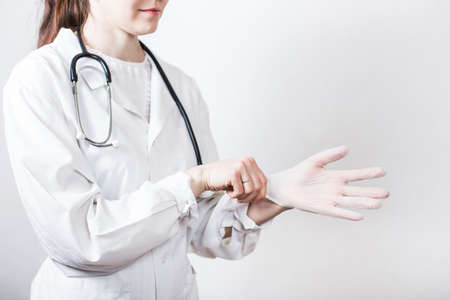 Portrait of young smiling woman brunette doctor in glasses and medical white coat with stethoscope putting on disposable medical gloves on white backgroundの写真素材