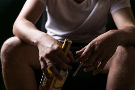alcohol and cigarette dependence. young sad man holding a cigarette and a bottle of alcohol in his handの写真素材