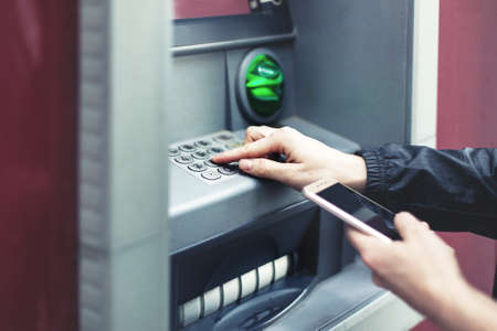 Closeup of female hands using smart phone while typing on ATM, bank machineの写真素材
