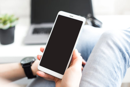Mockup image of woman's hands holding white mobile phone with blank screen on thigh in office