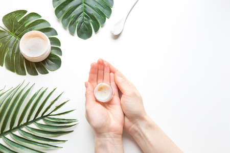 Flatlay. Woman putting nutritious cream on her hands on white background among jar of cosmetic cream, leaf palm branch. Final stage of manicure: woman uses moisturizer for the skin. Copy spaceの写真素材