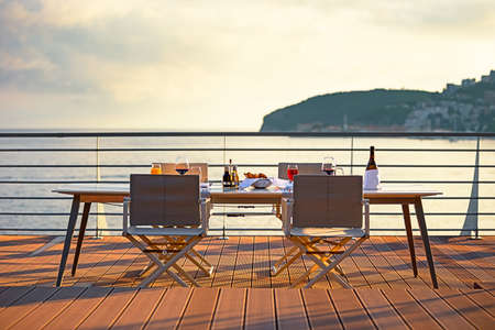 Table on the terrace at sunset overlooking the sea. Four chairs at the evening table with a gorgeous sea view. Bottle and glasses with red wine in sunset with sea view backgroundの写真素材