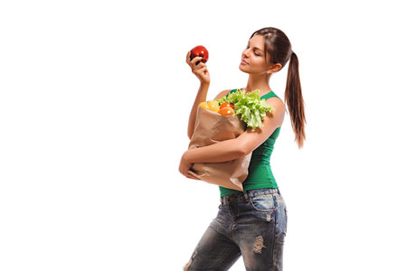 Girl holding bag with vegetables and fruits and looks at red apple in his hand. Woman with a paper...