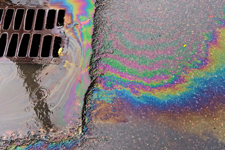 An oil slick against the backdrop of an asphalt road flows into a storm drain through a grate....