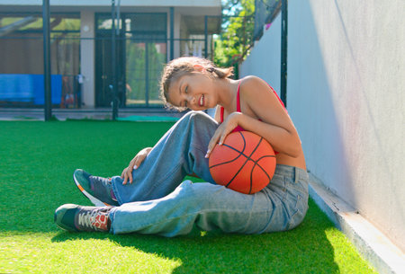 A girl sits with a basketball on the sports ground and laughs cheerfully.の写真素材
