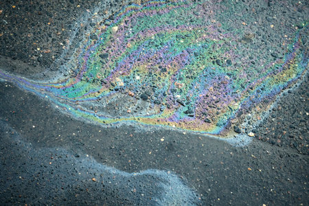 A vibrant oil slick creates a stunning display of colors on the pavement in an urban area, highlighting the intersection of nature and human activities during daylightの写真素材