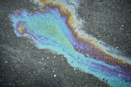 A vibrant oil spill spreads across the dark asphalt, revealing shades of blue, purple, and green. The unique patterns draw attention in the urban landscape, juxtaposed against the gray pavement.の写真素材