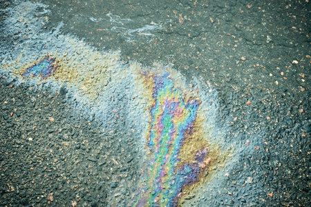 Colorful gasoline or oil spill creating a rainbow sheen on wet asphalt road, illustrating...