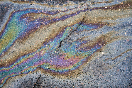 Oil slick creating a vibrant iridescent rainbow pattern on cracked asphalt, depicting environmental...