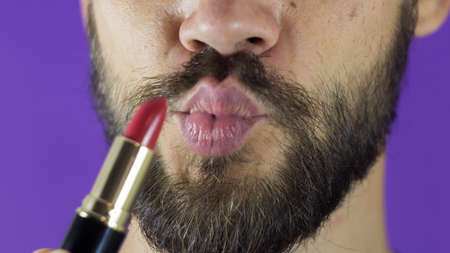 A young guy with a beard paints his lips with red lipstick. Close-up of a bearded man, he painted lips with bright lipstick. A bearded man puts red lipstick on his lips, smiles and smacks his lips.の写真素材