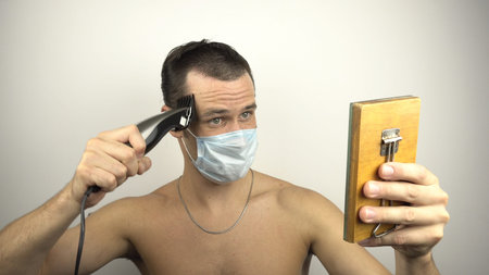 A young guy in a medical protective mask shaves his hair using a hair clipper while sitting at home...