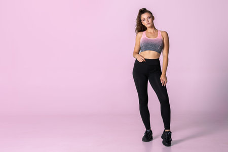 Athletic woman standing in fitness outfit with black leggings and a ponytail infront of a pink...