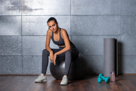 Female fitness trainer sits on a gymnastic ball with fitness equipment, dumbbells, mat and a bottle...
