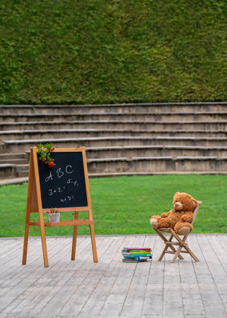 Back to school soon. In the open air there is a wooden board with inscriptions, and a yellow teddy bear sits on a small chair. There are books nearby. Postcard, congratulations, wishesの写真素材