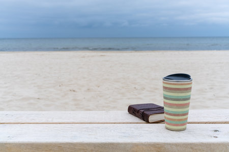 A glass of coffee and a notepad lie on a bench on a white sand beach. Against the backdrop of the sea. Congratulations, postcard, still lifeの写真素材