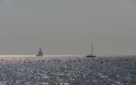 Two sailing yachts drifting in waters of the open sea against backdrop of picturesque sunset. Sunset sky is reflected on surface of water. Seascape, freedom, summer vacation.の写真素材
