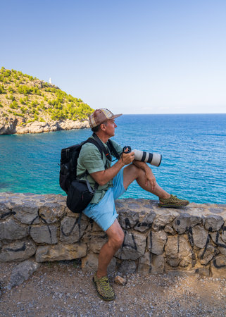 Travel photographer with professional camera sits on stone fence and enjoys sea view of Port de Soller of picturesque Mallorca. Photography, traveling, creative exploration.の写真素材