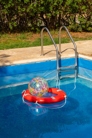 Colorful inflatable ball placed inside red lifebuoy floating in blue swimming pool. Symbol of child...