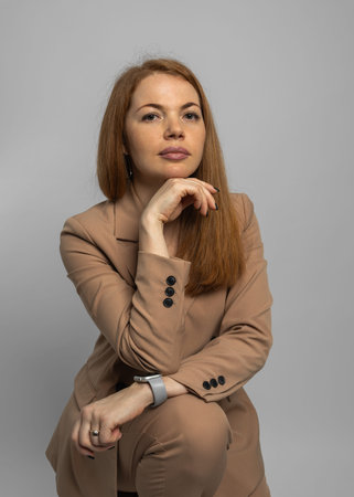 Stylish young woman with red hair in pantsuit posing on gray background, studio portrait. People...