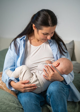 Mother breastfeeds her cute newborn baby sitting on her bed at home. Milk from mother's breast is a...