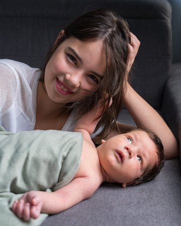 An older sister and her newborn little brother are lying on the sofa. The girl looks at the baby...
